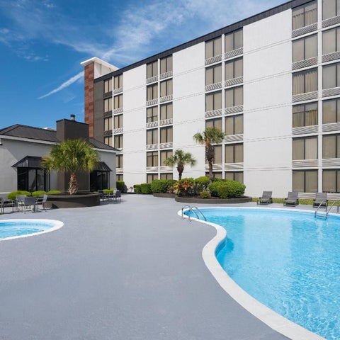 A large, curved swimming pool and jacuzzi are situated on a concrete patio in front of a tall, white hotel building.