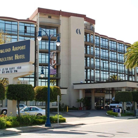 The Oakland Airport Executive Hotel features a modern facade with large windows, a sign, and lush landscaping.