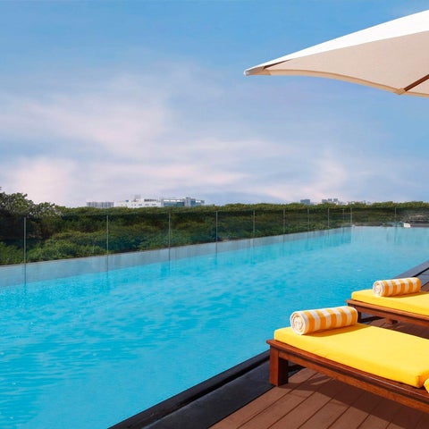 A vibrant turquoise infinity pool overlooks a lush green landscape with two yellow lounge chairs and a parasol.