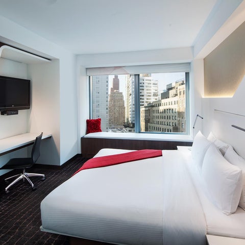 A modern hotel room features a king-size bed, a desk with a television, and a large window overlooking a cityscape.