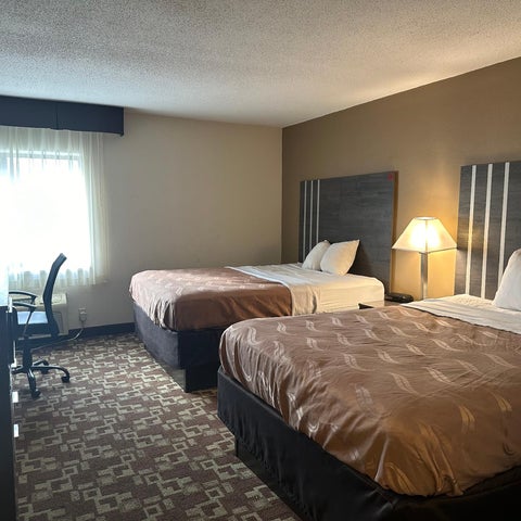 A hotel room features two queen beds with brown patterned comforters, bedside lamps, and a desk with a chair.