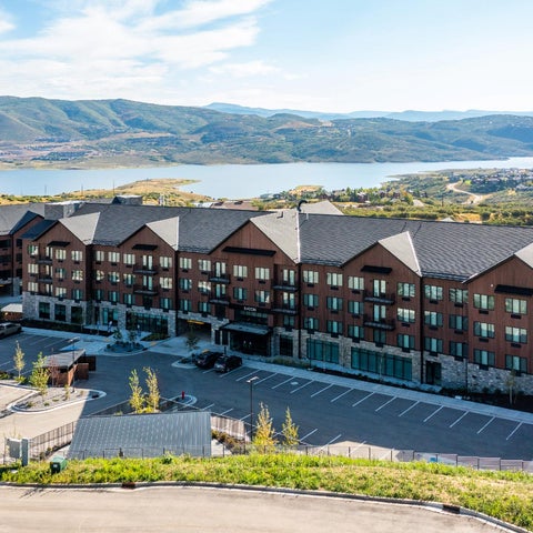A large, modern hotel building with a dark brown facade sits atop a hill overlooking a lake and distant mountains.