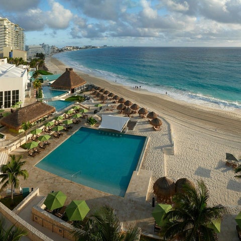 An aerial view showcases a resort with a turquoise pool, beach umbrellas, palm trees, and a sandy beach meeting the ocean.