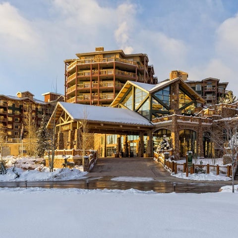 A large resort building with a covered entrance sits amidst snow-covered landscaping and a partially visible mountain backdrop.