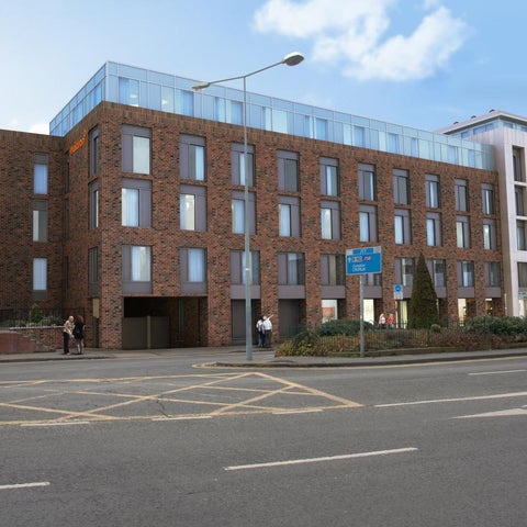 A modern brick building with a glass top floor and surrounding greenery stands on a street with pedestrians and traffic.
