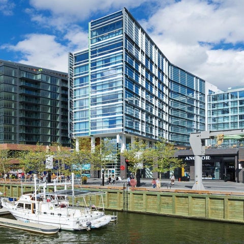 Modern glass and steel buildings line a waterfront promenade with boats docked and pedestrians strolling along the walkway.