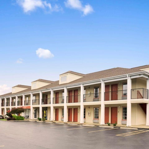 A two-story motel with a beige facade, white columns, and a parking lot under a partly cloudy sky.