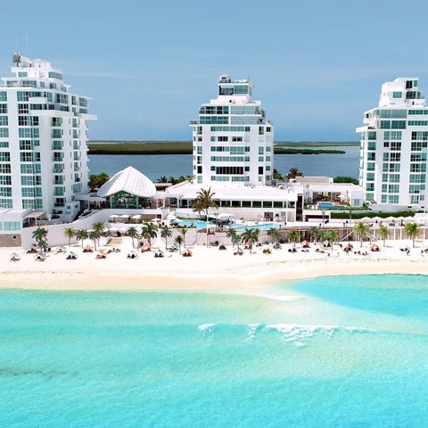 A wide aerial view showcases a pristine beach with turquoise water and several modern, white high-rise hotels.