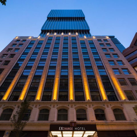 The Exchange Hotel's facade features a modern glass tower atop a classic stone base, illuminated with warm interior lighting.