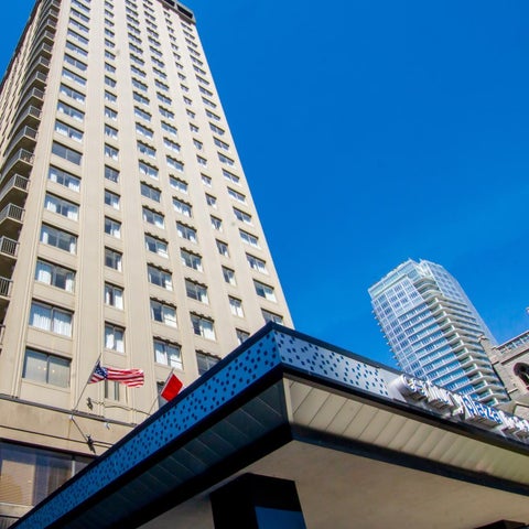 A tall, light-colored building rises against a blue sky, with other buildings and a rooftop structure in the foreground.