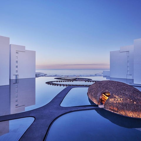 A modern hotel features a unique, egg-shaped structure in a reflecting pool surrounded by white buildings and a serene ocean view.