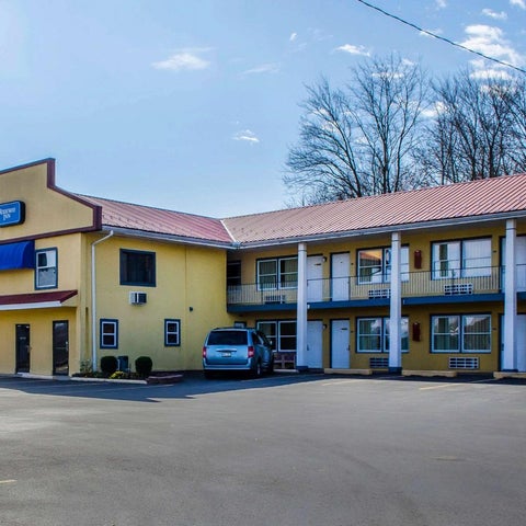 A two-story motel with a yellow facade and red roof sits alongside a parking lot under a partly cloudy sky.