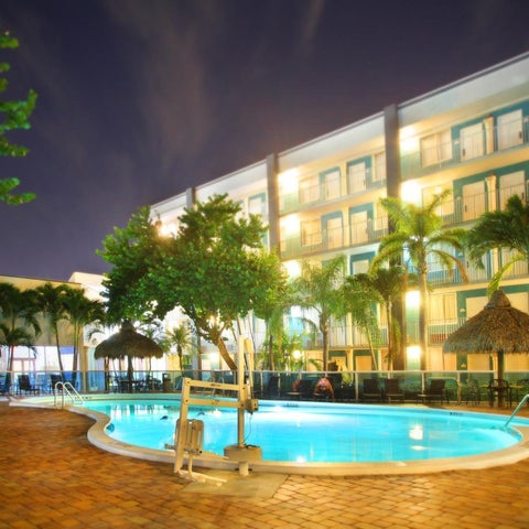 A brightly lit hotel pool with palm trees and thatched-roof cabanas surrounds a brick-paved patio area.
