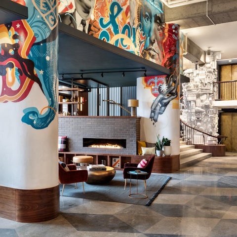 A modern hotel lobby features a vibrant, stylized mural on a pillar, a fireplace, and comfortable seating areas.
