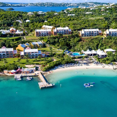 An aerial view showcases a resort with multiple buildings, a sandy beach, a dock, and a pool area with lounge chairs.