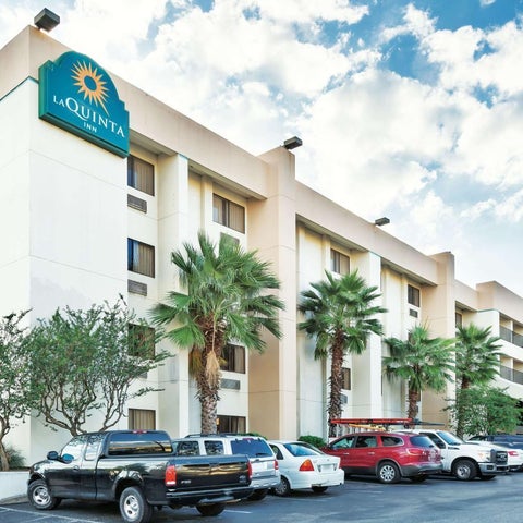 A La Quinta Inn building features a light-colored facade with palm trees, parking, and several vehicles in the foreground.