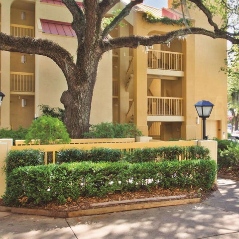 A multi-story building with balconies is framed by a large tree and manicured landscaping with decorative lighting.