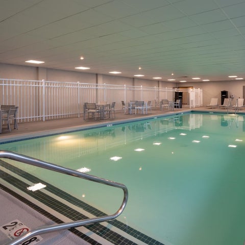 An indoor pool with a light blue hue is surrounded by lounge chairs and tables in a well-lit space.
