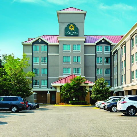 A La Quinta Inn building features a multi-story facade with a prominent logo, parking lot, and surrounding greenery.