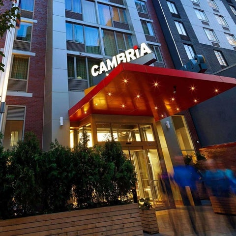 The Cambria Hotel entrance features a red awning, glass doors, and lush greenery in front of a brick building.