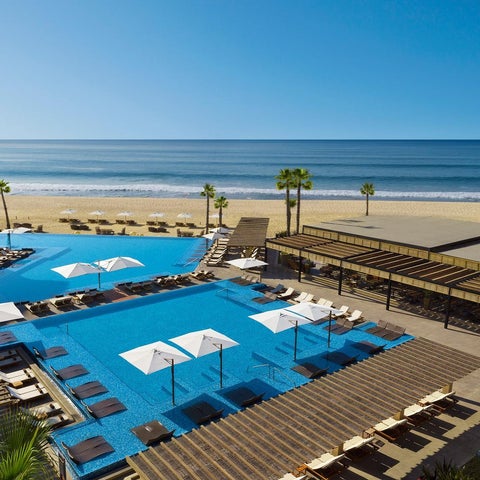 An aerial view showcases a large, turquoise pool with lounge chairs and umbrellas, adjacent to a sandy beach and palm trees.