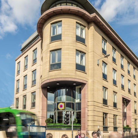 A modern, multi-story building with a curved facade and a bus driving past pedestrians on a sunny day.