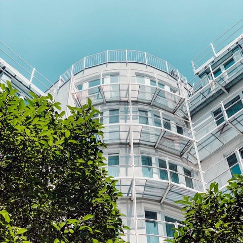 A modern, white building with rounded corners and balconies is partially framed by green foliage in the foreground.