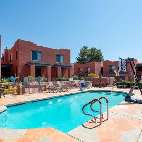 A bright blue pool surrounded by lounge chairs, tables, and a basketball hoop sits in front of a multi-building complex.