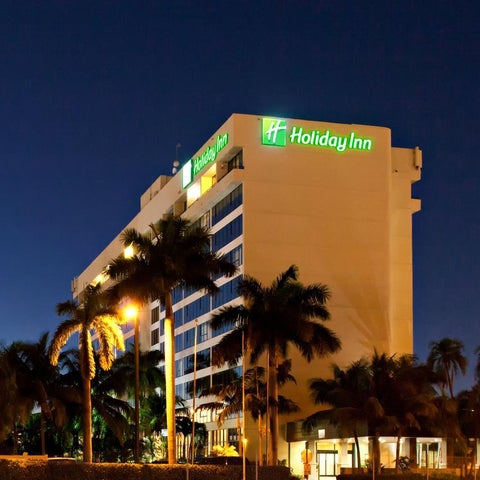 A Holiday Inn hotel building is illuminated at night, featuring palm trees and outdoor lighting.