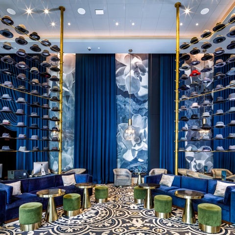 A modern lobby features a blue velvet sofa, circular tables, and a display of hats suspended from gold poles against a patterned backdrop.