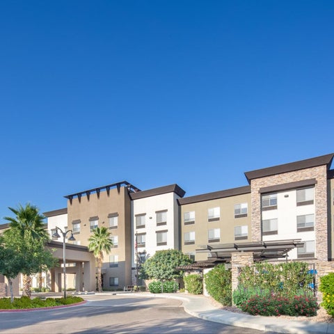 A multi-story hotel building with a covered entrance and landscaping is seen against a clear blue sky.