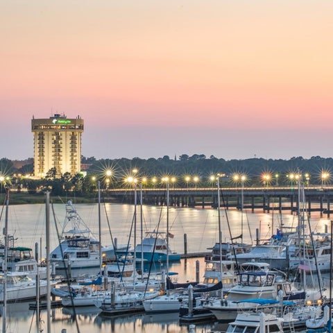 A waterfront view showcases a hotel with a tall tower, a marina filled with boats, and a bridge under a colorful sunset sky.