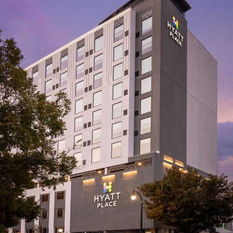 A modern Hyatt Place hotel building is shown with a gray and white facade under a colorful twilight sky.