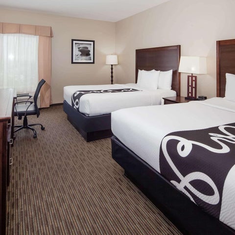 A hotel room features two queen beds with dark bedding and bedside lamps, a desk, and a window with curtains.