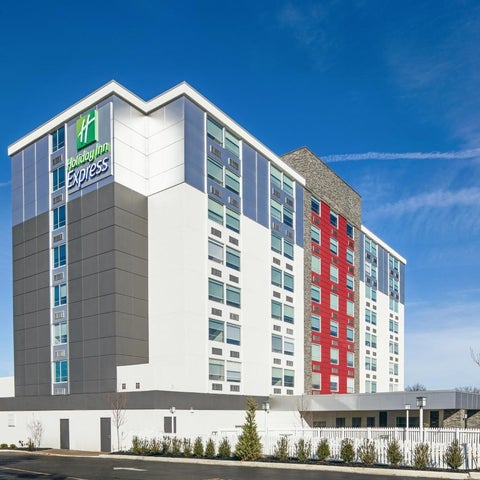 A modern, multi-story Holiday Inn Express hotel building stands against a bright blue sky with a clear view of the surrounding area.