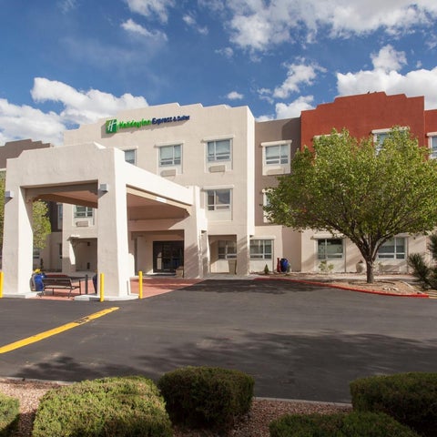 A multistory hotel with a covered entrance and parking lot is seen on a sunny day with scattered clouds.