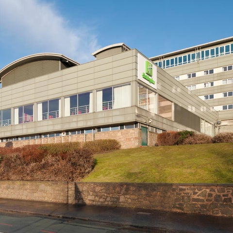A modern Holiday Inn hotel building sits on a slight hill with a stone retaining wall and landscaping in front.