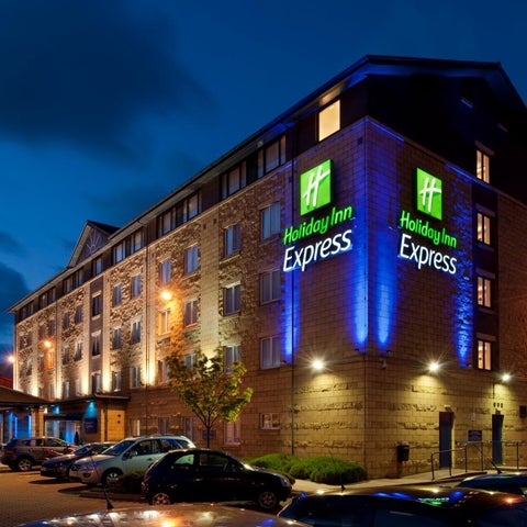 A Holiday Inn Express hotel building is illuminated at night with blue and yellow lighting, featuring a parking lot and surrounding cityscape.