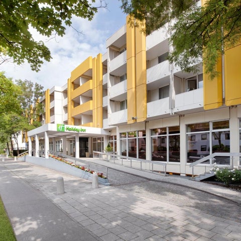 A multi-story Holiday Inn hotel building with a brick facade and yellow accents stands alongside a sidewalk and lush greenery.