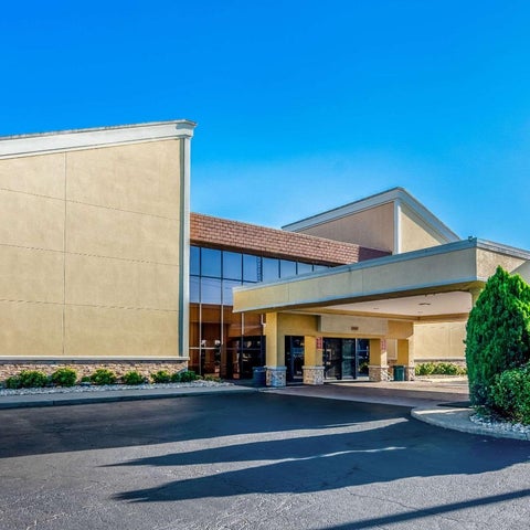 A beige hotel building with a covered entrance, landscaping, and a sign visible against a clear blue sky.