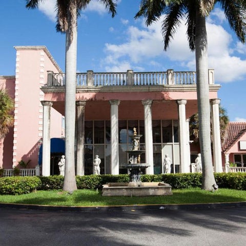 A pink-colored hotel building with classical columns, statues, and a fountain sits on a circular driveway.