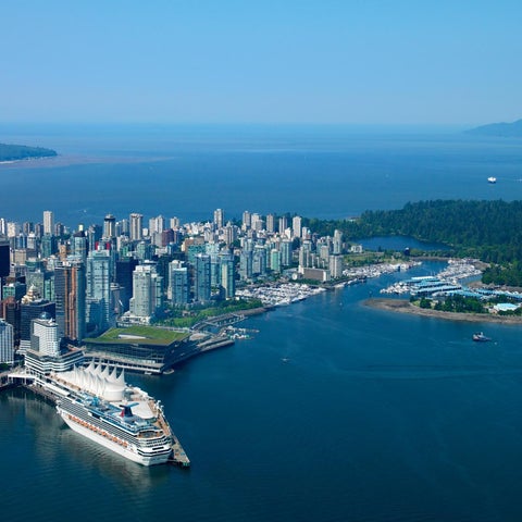 An aerial view showcases Vancouver's skyline, waterfront, cruise ship terminal, and lush green landscape against a clear blue sky.
