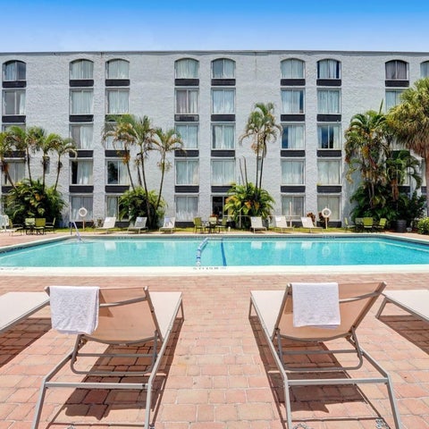 A rectangular swimming pool with lounge chairs and towels surrounds a large, white hotel building with palm trees.