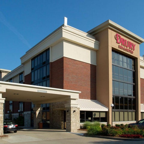 A multi-story Drury hotel building with a covered entrance and parking area, featuring a brick and stucco facade.