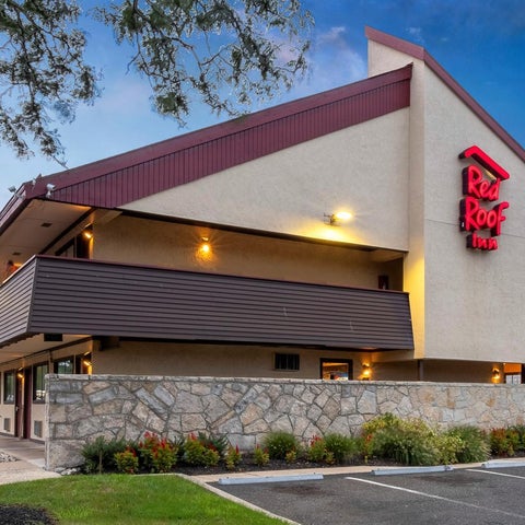 A Red Roof Inn building features a red roof, beige facade, and a stone base with landscaping and parking in front.