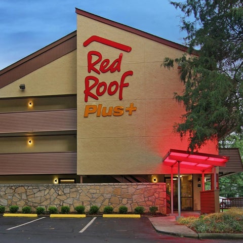 A Red Roof Plus hotel building is shown with a tan facade, red signage, and a stone wall in front of the entrance.