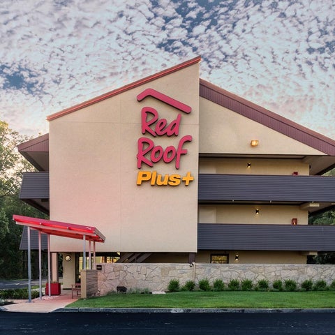 A Red Roof Plus hotel building is shown with its signage and landscaping against a cloudy sky.