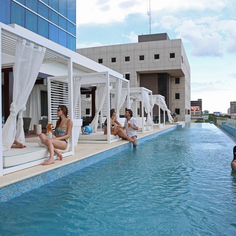 Guests relax and swim in a long, rectangular pool with cabanas and a modern building backdrop.