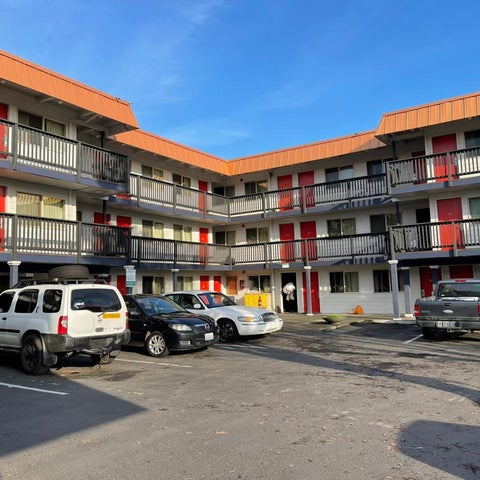 A multi-story motel with red and white facades features a parking lot with several cars parked in front.