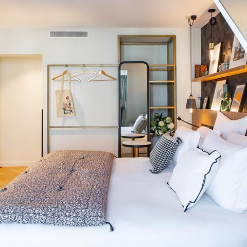 A modern hotel room features a large bed with patterned bedding, a desk, and stylish decor including a mirror and shelving.